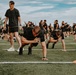 A large group of individuals dressed in matching black t-shirts and shorts are participating in an organized physical training session on an artificial turf field. Several participants in the foreground are performing push-ups or a push-up variation, while one standing individual appears to be an instructor or supervisor holding a medicine ball. The background shows dozens more participants engaged in various exercises, suggesting a structured group fitness or military-style PT event.