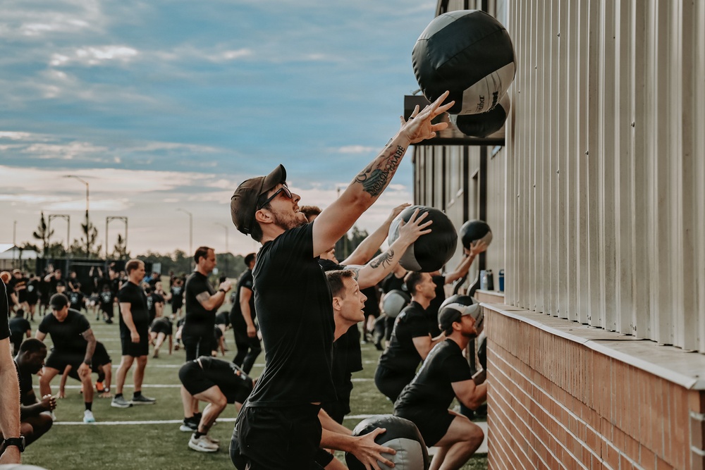 A large group of people dressed uniformly in black athletic clothing are performing a synchronized outdoor fitness workout. The foreground shows participants doing wall ball exercises, throwing medicine balls against the side of a building. The event appears to be a large-scale group fitness class or military/tactical training event held on a sports field.