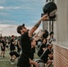 A large group of people dressed uniformly in black athletic clothing are performing a synchronized outdoor fitness workout. The foreground shows participants doing wall ball exercises, throwing medicine balls against the side of a building. The event appears to be a large-scale group fitness class or military/tactical training event held on a sports field.
