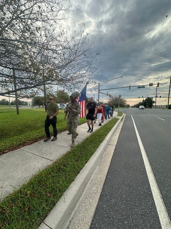 A group of people are walking along a sidewalk beside a multi-lane road under a dramatic, overcast sky. One individual in military camouflage uniform is prominently carrying a large American flag. The scene appears to be an organized patriotic march or ruck walk event.