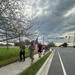 A group of people are walking along a sidewalk beside a multi-lane road under a dramatic, overcast sky. One individual in military camouflage uniform is prominently carrying a large American flag. The scene appears to be an organized patriotic march or ruck walk event.