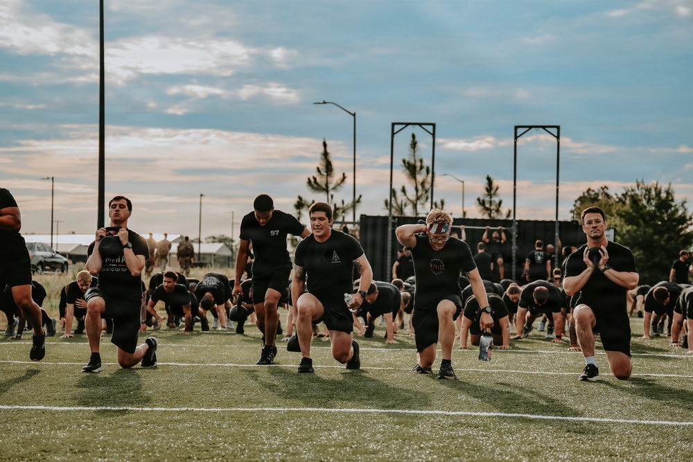 A large group of individuals dressed in black athletic wear are performing a synchronized outdoor fitness workout on a turf field. The foreground shows participants doing lunges while holding weighted objects, with many others in the background performing push-ups or similar ground exercises. The setting appears to be a military or organized group physical training event, likely at dusk given the overcast sky and lighting conditions.