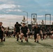 A large group of individuals dressed in black athletic wear are performing a synchronized outdoor fitness workout on a turf field. The foreground shows participants doing lunges while holding weighted objects, with many others in the background performing push-ups or similar ground exercises. The setting appears to be a military or organized group physical training event, likely at dusk given the overcast sky and lighting conditions.