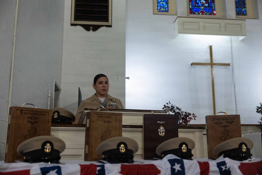 A U.S. Navy enlisted service member in khaki uniform speaks at a podium during a formal military ceremony held inside a chapel. Several wooden plaques and Navy chief petty officer covers (hats) are displayed on a table in the foreground, along with what appears to be an American flag. The setting, cross, and stained glass windows confirm a military chapel environment, consistent with a chief petty officer pinning or advancement ceremony.