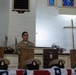A U.S. Navy enlisted service member in khaki uniform speaks at a podium during a formal military ceremony held inside a chapel. Several wooden plaques and Navy chief petty officer covers (hats) are displayed on a table in the foreground, along with what appears to be an American flag. The setting, cross, and stained glass windows confirm a military chapel environment, consistent with a chief petty officer pinning or advancement ceremony.