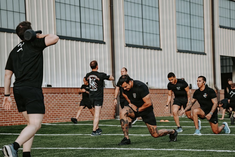 A group of individuals dressed in black athletic wear are performing physical training exercises on an outdoor turf field. The session appears to be a structured group workout, with participants performing lunges and other functional movements. A large industrial or hangar-style building with brick and corrugated metal siding serves as the backdrop.