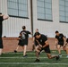 A group of individuals dressed in black athletic wear are performing physical training exercises on an outdoor turf field. The session appears to be a structured group workout, with participants performing lunges and other functional movements. A large industrial or hangar-style building with brick and corrugated metal siding serves as the backdrop.