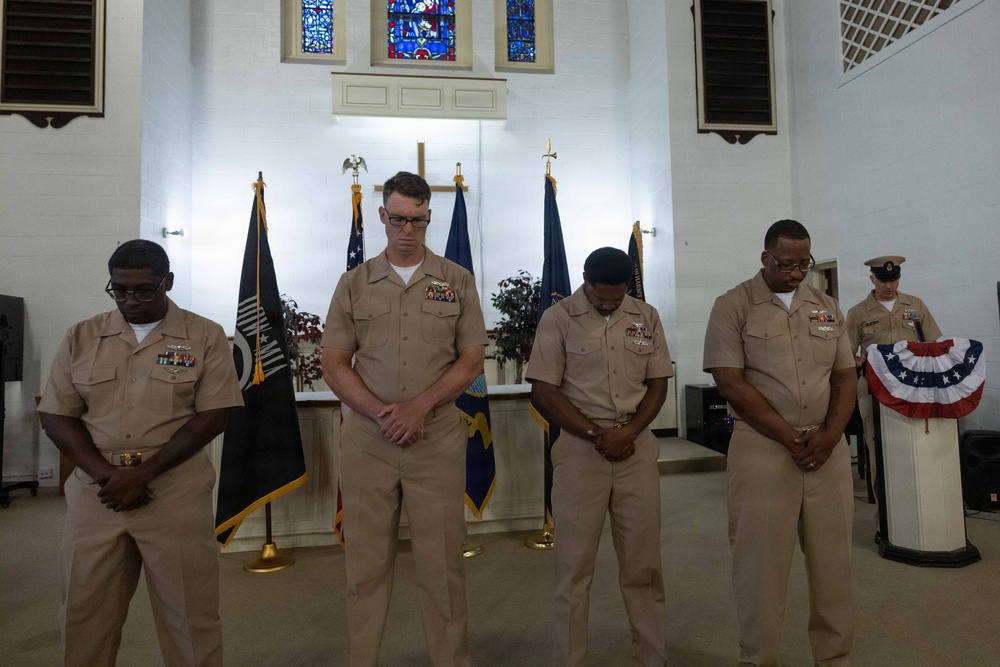 Four U.S. Navy personnel in tan service uniforms stand with heads bowed in what appears to be a prayer or moment of silence during a formal military ceremony. The setting is a chapel or ceremonial hall, identifiable by stained glass windows, a cross, and multiple flags including a POW/MIA flag. A fifth service member stands at a podium in the background.