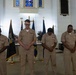 Four U.S. Navy personnel in tan service uniforms stand with heads bowed in what appears to be a prayer or moment of silence during a formal military ceremony. The setting is a chapel or ceremonial hall, identifiable by stained glass windows, a cross, and multiple flags including a POW/MIA flag. A fifth service member stands at a podium in the background.