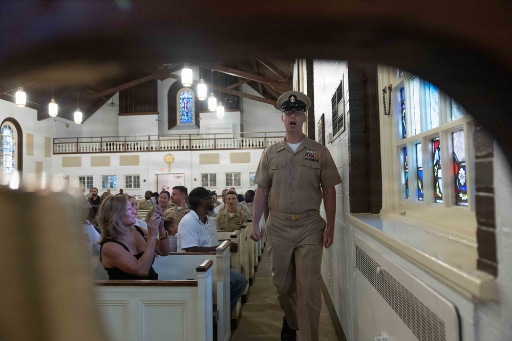 A U.S. Navy officer in khaki service uniform walks down the side aisle of a chapel or church, appearing to speak or call out to the congregation. The pews are filled with a mix of uniformed Navy personnel and civilians. The setting appears to be a military chapel, evidenced by the traditional church architecture including stained glass windows, wooden pews, and an upper balcony.