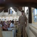 A U.S. Navy officer in khaki service uniform walks down the side aisle of a chapel or church, appearing to speak or call out to the congregation. The pews are filled with a mix of uniformed Navy personnel and civilians. The setting appears to be a military chapel, evidenced by the traditional church architecture including stained glass windows, wooden pews, and an upper balcony.