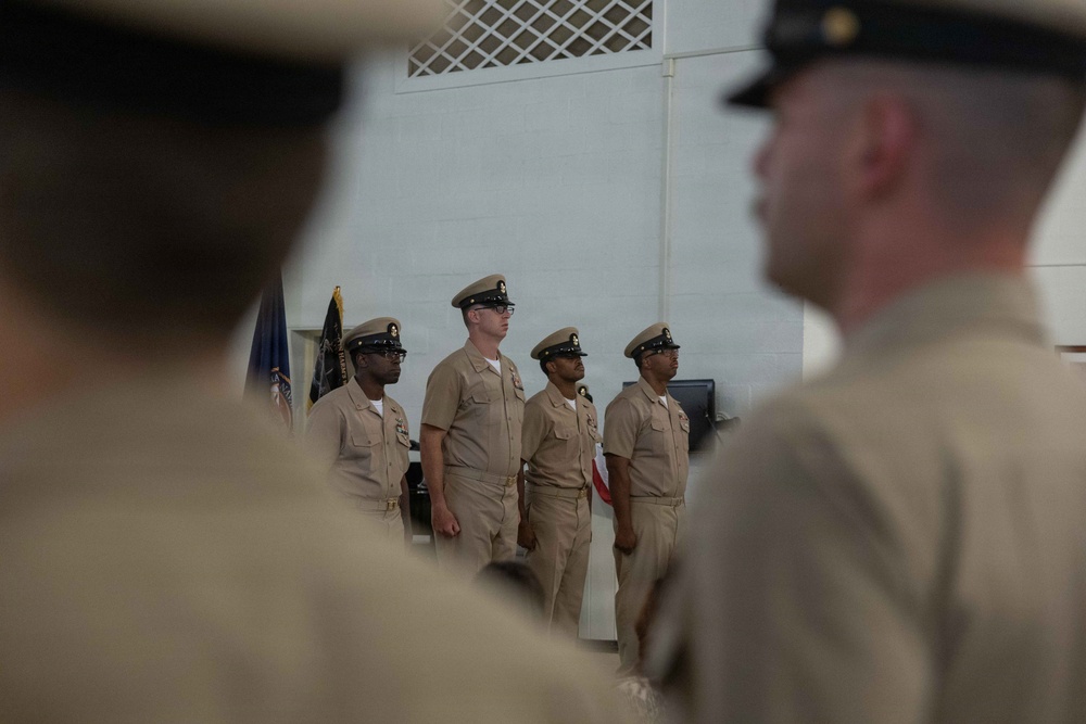 A formal military ceremony featuring personnel in U.S. Navy Service Khaki uniforms standing at attention in an indoor facility. Four sailors are visible in the mid-ground standing in a line, while two additional uniformed personnel are partially visible in the foreground, blurred due to depth of field. A U.S. Navy flag or guidon is partially visible in the background.
