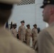 A formal military ceremony featuring personnel in U.S. Navy Service Khaki uniforms standing at attention in an indoor facility. Four sailors are visible in the mid-ground standing in a line, while two additional uniformed personnel are partially visible in the foreground, blurred due to depth of field. A U.S. Navy flag or guidon is partially visible in the background.