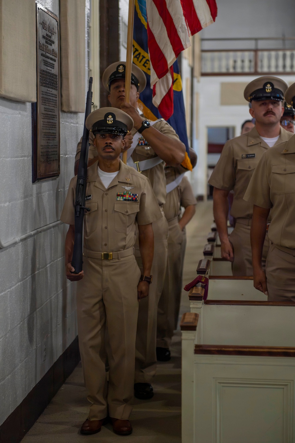 A U.S. Navy color guard ceremony is taking place inside what appears to be a chapel or ceremonial hall. Navy Chief Petty Officers in khaki service uniforms are participating in a formal procession, with one sailor carrying a rifle at port arms and another bearing the U.S. flag alongside what appears to be a state flag. A commemorative plaque is visible on the wall to the left.