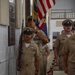 A U.S. Navy color guard ceremony is taking place inside what appears to be a chapel or ceremonial hall. Navy Chief Petty Officers in khaki service uniforms are participating in a formal procession, with one sailor carrying a rifle at port arms and another bearing the U.S. flag alongside what appears to be a state flag. A commemorative plaque is visible on the wall to the left.