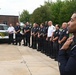 A group of approximately 13-15 law enforcement and emergency personnel are standing in a line outdoors, appearing to be at attention or in formation. A Military Police vehicle (SUV) is visible in the background along with a brick building and trees. The scene suggests a formal gathering, ceremony, or briefing at what appears to be a military installation.