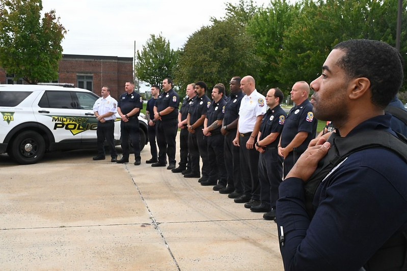 A group of approximately 13-15 law enforcement and emergency personnel are standing in a line outdoors, appearing to be at attention or in formation. A Military Police vehicle (SUV) is visible in the background along with a brick building and trees. The scene suggests a formal gathering, ceremony, or briefing at what appears to be a military installation.