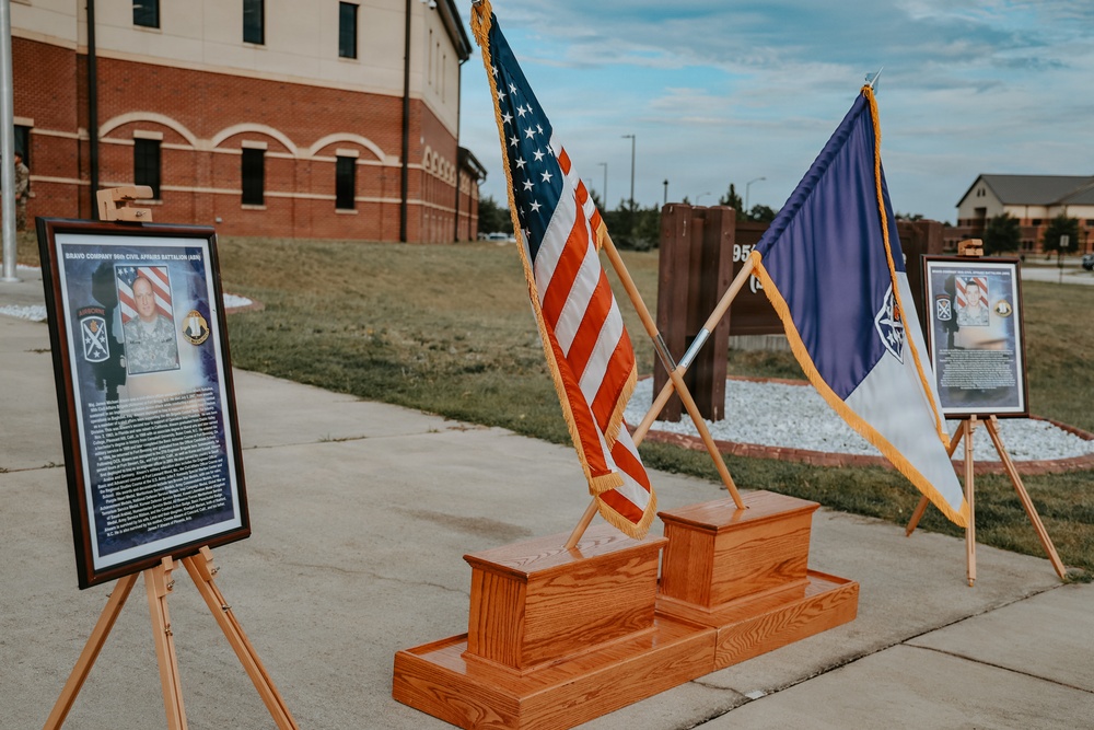 A military memorial or dedication ceremony display is set up outdoors on what appears to be a military installation. Two framed commemorative plaques on easels flank a wooden stepped podium holding crossed flags — the United States flag and what appears to be a unit or organizational flag. The setting includes brick military buildings in the background, consistent with a U.S. Army post.