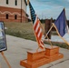 A military memorial or dedication ceremony display is set up outdoors on what appears to be a military installation. Two framed commemorative plaques on easels flank a wooden stepped podium holding crossed flags — the United States flag and what appears to be a unit or organizational flag. The setting includes brick military buildings in the background, consistent with a U.S. Army post.