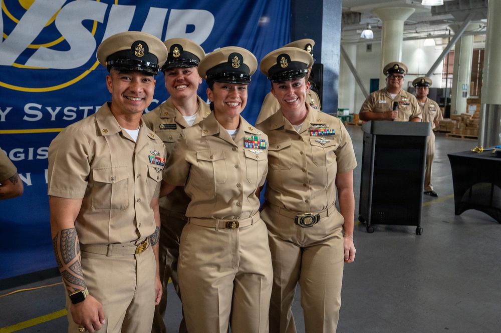 Four U.S. Navy personnel in Service Khaki uniforms pose together smiling at what appears to be a military ceremony or event. They are standing in front of a blue banner partially reading 'SUR' and 'Logistic System Command' (NAVSUP). In the background, two additional Navy personnel stand near a podium, suggesting an official ceremony is taking place.
