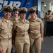 Four U.S. Navy personnel in Service Khaki uniforms pose together smiling at what appears to be a military ceremony or event. They are standing in front of a blue banner partially reading 'SUR' and 'Logistic System Command' (NAVSUP). In the background, two additional Navy personnel stand near a podium, suggesting an official ceremony is taking place.
