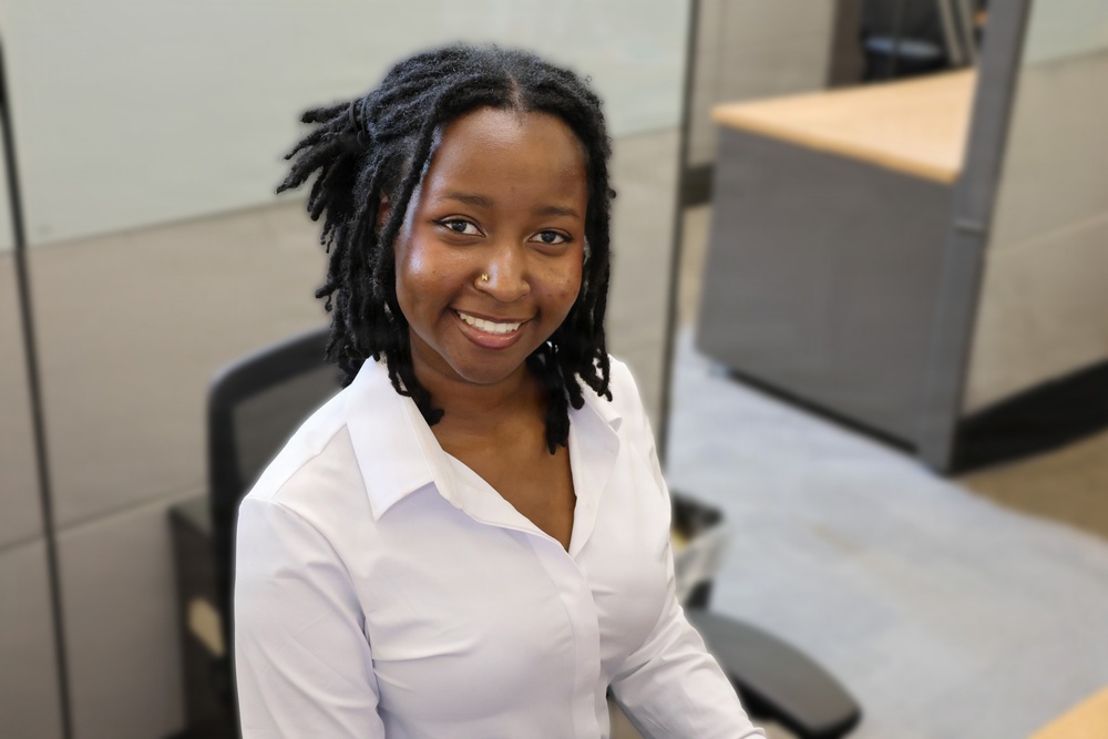 A young woman is seated in an office cubicle environment, smiling warmly at the camera. She is wearing a white button-up shirt and has short dreadlocks, with a small nose piercing visible. The photo appears to be a professional or workplace portrait taken from a slightly elevated angle.