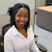 A young woman is seated in an office cubicle environment, smiling warmly at the camera. She is wearing a white button-up shirt and has short dreadlocks, with a small nose piercing visible. The photo appears to be a professional or workplace portrait taken from a slightly elevated angle.