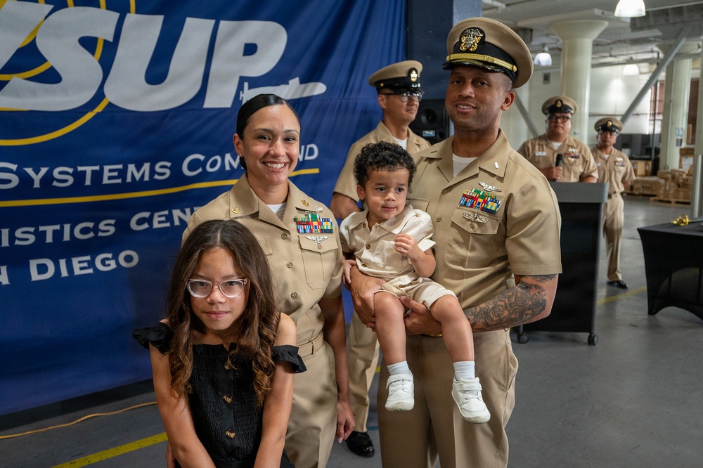 A U.S. Navy family poses for a photo at what appears to be a military ceremony or promotion event at the Fleet Logistics Center in San Diego. Two Navy personnel in Service Khaki uniforms stand together holding a toddler, accompanied by a young girl in a black dress. Additional Navy personnel are visible in the background near a blue banner.