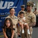 A U.S. Navy family poses for a photo at what appears to be a military ceremony or promotion event at the Fleet Logistics Center in San Diego. Two Navy personnel in Service Khaki uniforms stand together holding a toddler, accompanied by a young girl in a black dress. Additional Navy personnel are visible in the background near a blue banner.