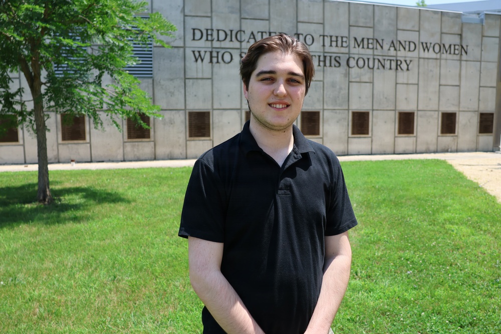 A young man wearing a black polo shirt stands on a green lawn in front of a large institutional building. The building features an engraved dedication inscription reading 'DEDICATED TO THE MEN AND WOMEN WHO S[ERVED] THIS COUNTRY,' suggesting a memorial or veterans-related facility. The photo appears to be taken on a sunny day with clear outdoor lighting.
