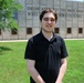 A young man wearing a black polo shirt stands on a green lawn in front of a large institutional building. The building features an engraved dedication inscription reading 'DEDICATED TO THE MEN AND WOMEN WHO S[ERVED] THIS COUNTRY,' suggesting a memorial or veterans-related facility. The photo appears to be taken on a sunny day with clear outdoor lighting.