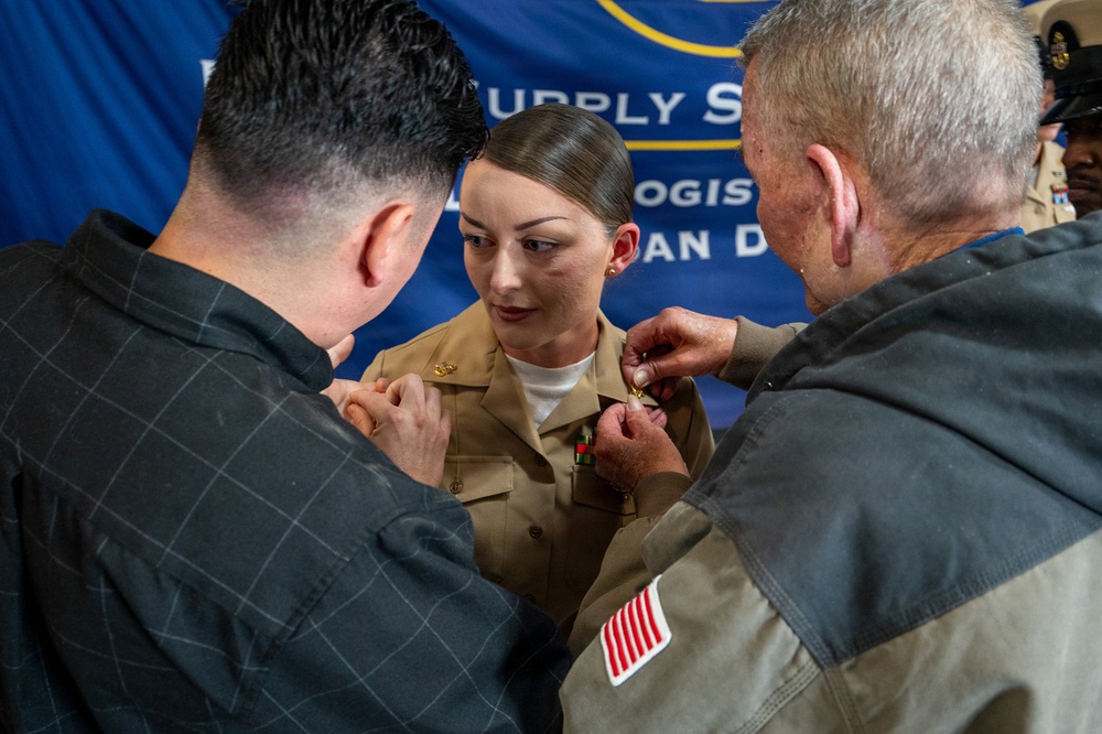A female military service member in a tan/khaki uniform is having insignia or medals pinned onto her uniform by two civilians during what appears to be a promotion or award ceremony. The event is taking place in front of a blue banner that partially reads 'Supply' and 'Logistics' and 'San D' (likely San Diego). Two individuals, one on each side, are simultaneously pinning items to her uniform chest area.