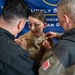 A female military service member in a tan/khaki uniform is having insignia or medals pinned onto her uniform by two civilians during what appears to be a promotion or award ceremony. The event is taking place in front of a blue banner that partially reads 'Supply' and 'Logistics' and 'San D' (likely San Diego). Two individuals, one on each side, are simultaneously pinning items to her uniform chest area.