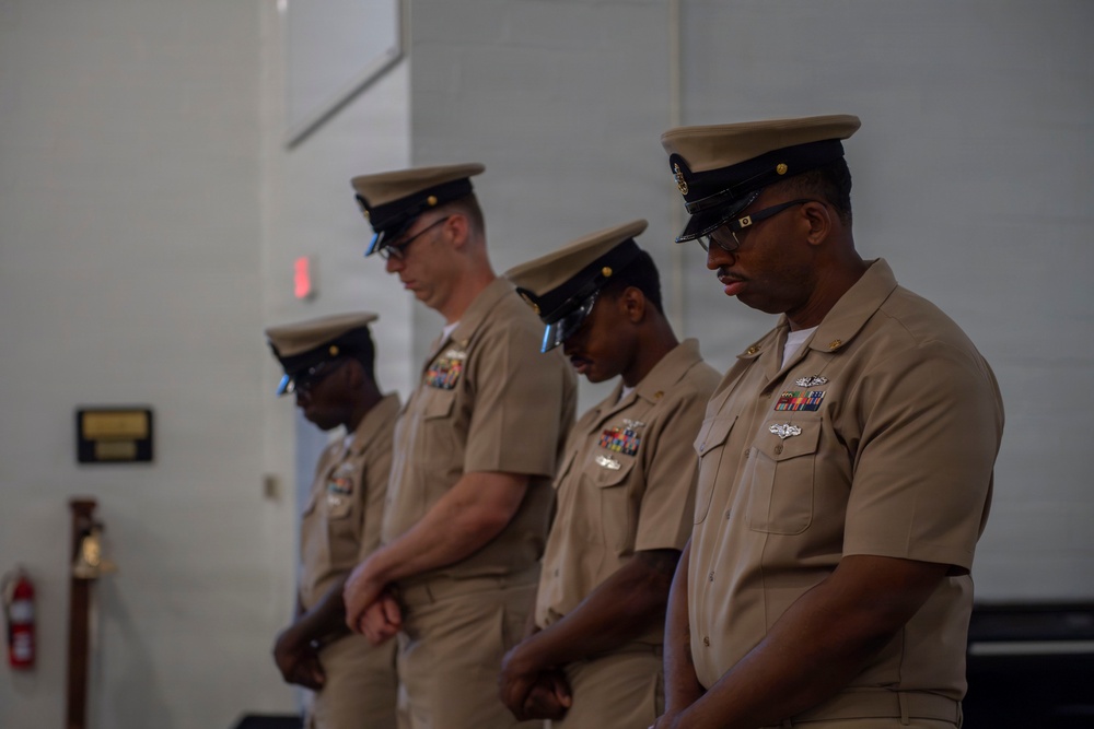 Four U.S. Navy personnel in tan/khaki service uniforms are standing in a row with heads bowed, suggesting a moment of prayer or solemn ceremony. They are indoors, likely in a military facility or ceremonial hall. All individuals are wearing combination covers (caps) and display various ribbons and insignia on their uniforms.