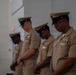Four U.S. Navy personnel in tan/khaki service uniforms are standing in a row with heads bowed, suggesting a moment of prayer or solemn ceremony. They are indoors, likely in a military facility or ceremonial hall. All individuals are wearing combination covers (caps) and display various ribbons and insignia on their uniforms.