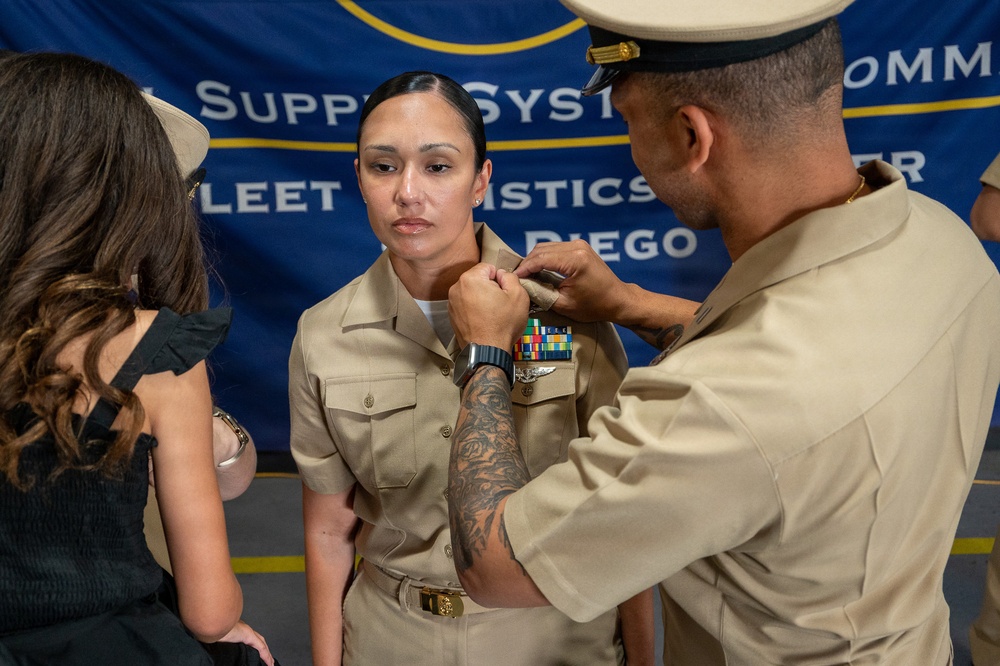 A U.S. Navy promotion or pinning ceremony taking place in front of a Fleet Logistics Center San Diego banner. A male sailor in khaki uniform with a cover (hat) is pinning insignia or a device onto the collar of a female sailor in khaki service uniform. A civilian woman in a black dress observes from the left side.