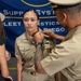 A U.S. Navy promotion or pinning ceremony taking place in front of a Fleet Logistics Center San Diego banner. A male sailor in khaki uniform with a cover (hat) is pinning insignia or a device onto the collar of a female sailor in khaki service uniform. A civilian woman in a black dress observes from the left side.
