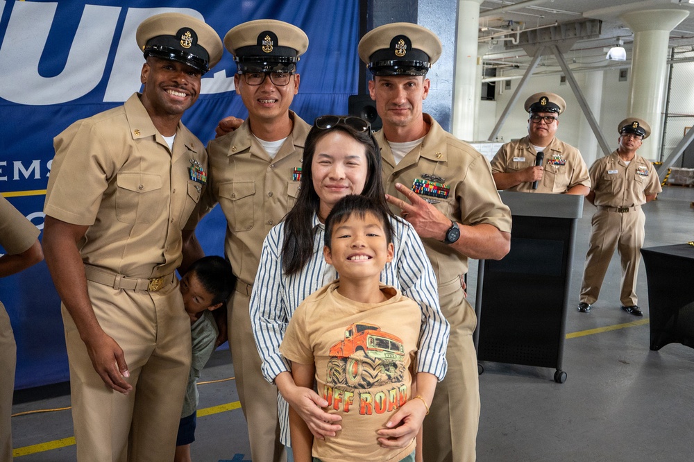 A group photo at what appears to be a U.S. Navy ceremony or event, featuring three Navy Chief Petty Officers in tan service uniforms posing with a woman and a young boy. The setting appears to be inside a naval vessel or military facility, with a blue banner visible in the background. Two additional Navy personnel are visible in the background near a podium.