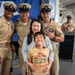 A group photo at what appears to be a U.S. Navy ceremony or event, featuring three Navy Chief Petty Officers in tan service uniforms posing with a woman and a young boy. The setting appears to be inside a naval vessel or military facility, with a blue banner visible in the background. Two additional Navy personnel are visible in the background near a podium.