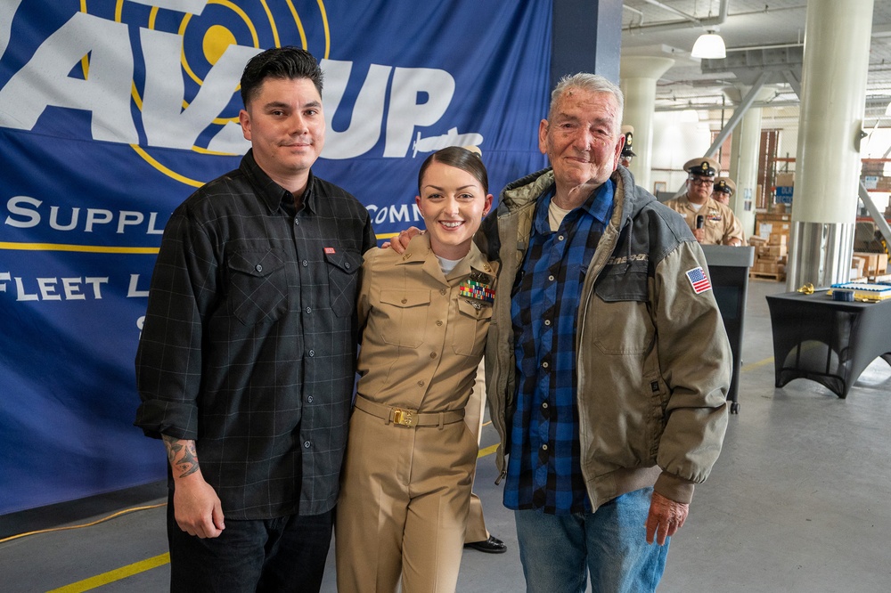 Three people pose together for a photo at what appears to be a military ceremony or event. The central figure is a woman in U.S. Navy dress uniform with multiple service ribbons/medals, flanked by a younger man in casual dark clothing and an older man in a jacket with an American flag patch. A blue banner in the background references a Navy supply or fleet logistics command.