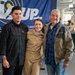 Three people pose together for a photo at what appears to be a military ceremony or event. The central figure is a woman in U.S. Navy dress uniform with multiple service ribbons/medals, flanked by a younger man in casual dark clothing and an older man in a jacket with an American flag patch. A blue banner in the background references a Navy supply or fleet logistics command.