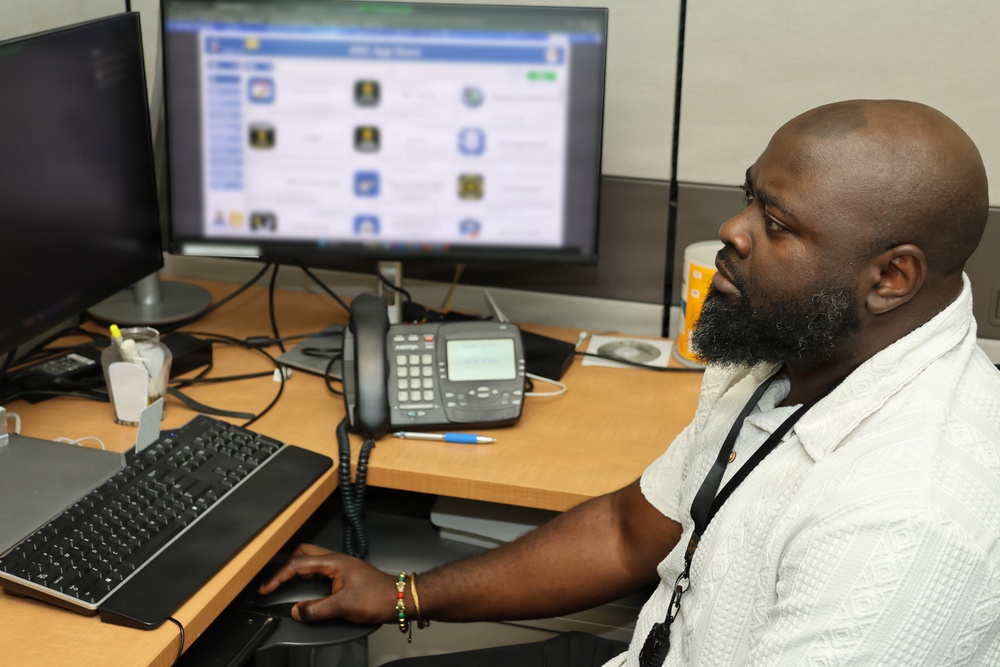 A bald man with a beard is seated at an office workstation, looking at a computer monitor displaying what appears to be a web page or app store interface. He is wearing a white shirt and a lanyard with an ID badge, and is using a mouse at his desk. The workspace includes dual monitors, a desk phone, keyboard, and various office items.