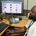 A bald man with a beard is seated at an office workstation, looking at a computer monitor displaying what appears to be a web page or app store interface. He is wearing a white shirt and a lanyard with an ID badge, and is using a mouse at his desk. The workspace includes dual monitors, a desk phone, keyboard, and various office items.
