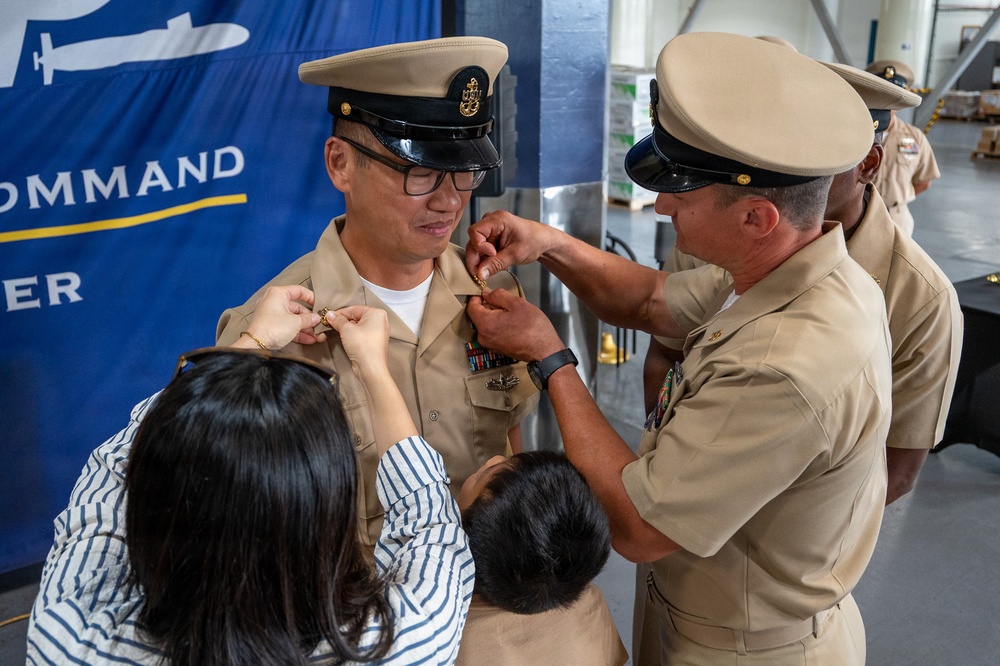 A U.S. Navy promotion or pinning ceremony is taking place in what appears to be a military facility. A sailor in khaki dress uniform is being pinned simultaneously by a civilian woman on his left shoulder and a senior enlisted sailor on his right collar area, while a child stands nearby. A blue command banner is visible in the background.