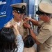 A U.S. Navy promotion or pinning ceremony is taking place in what appears to be a military facility. A sailor in khaki dress uniform is being pinned simultaneously by a civilian woman on his left shoulder and a senior enlisted sailor on his right collar area, while a child stands nearby. A blue command banner is visible in the background.