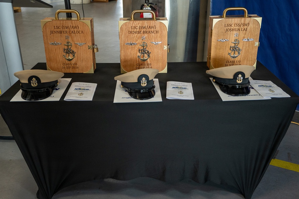 A ceremonial display table covered in black cloth holds three personalized wooden keepsake boxes and three U.S. Navy Chief Petty Officer covers (hats) alongside printed documents. The wooden boxes are engraved with individual names, ranks, warfare qualifications, and the date '16 SEP 2026, Class 133,' indicating a Navy Chief Petty Officer pinning or advancement ceremony. The items belong to LSC (SW/AW) Jennifer Caloca, LSC (SW/AW) Denise Branch, and LSSC (SS/SW) Joshua Lai.