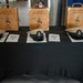 A ceremonial display table covered in black cloth holds three personalized wooden keepsake boxes and three U.S. Navy Chief Petty Officer covers (hats) alongside printed documents. The wooden boxes are engraved with individual names, ranks, warfare qualifications, and the date '16 SEP 2026, Class 133,' indicating a Navy Chief Petty Officer pinning or advancement ceremony. The items belong to LSC (SW/AW) Jennifer Caloca, LSC (SW/AW) Denise Branch, and LSSC (SS/SW) Joshua Lai.