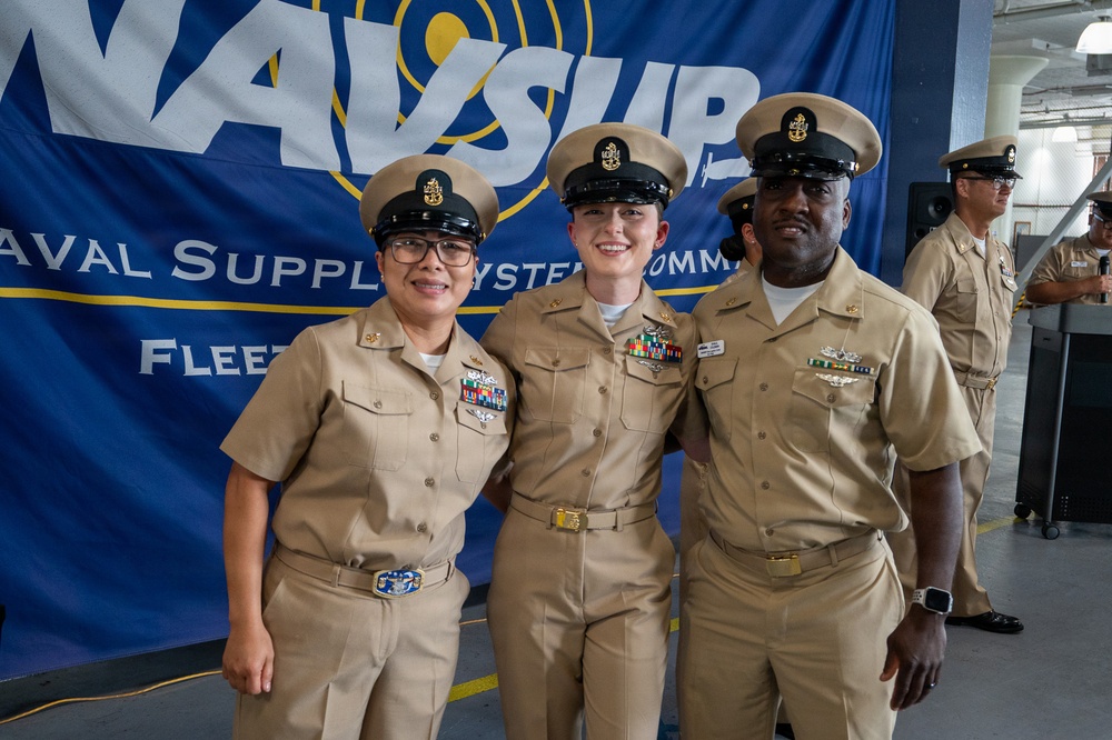 Three U.S. Navy personnel in khaki service uniforms pose together in front of a Naval Supply Systems Command (NAVSUP) banner. The central figure appears to be the focus of a ceremony or promotion event, wearing multiple ribbons and decorations. A fourth service member is visible in the background near audio equipment.