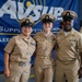 Three U.S. Navy personnel in khaki service uniforms pose together in front of a Naval Supply Systems Command (NAVSUP) banner. The central figure appears to be the focus of a ceremony or promotion event, wearing multiple ribbons and decorations. A fourth service member is visible in the background near audio equipment.
