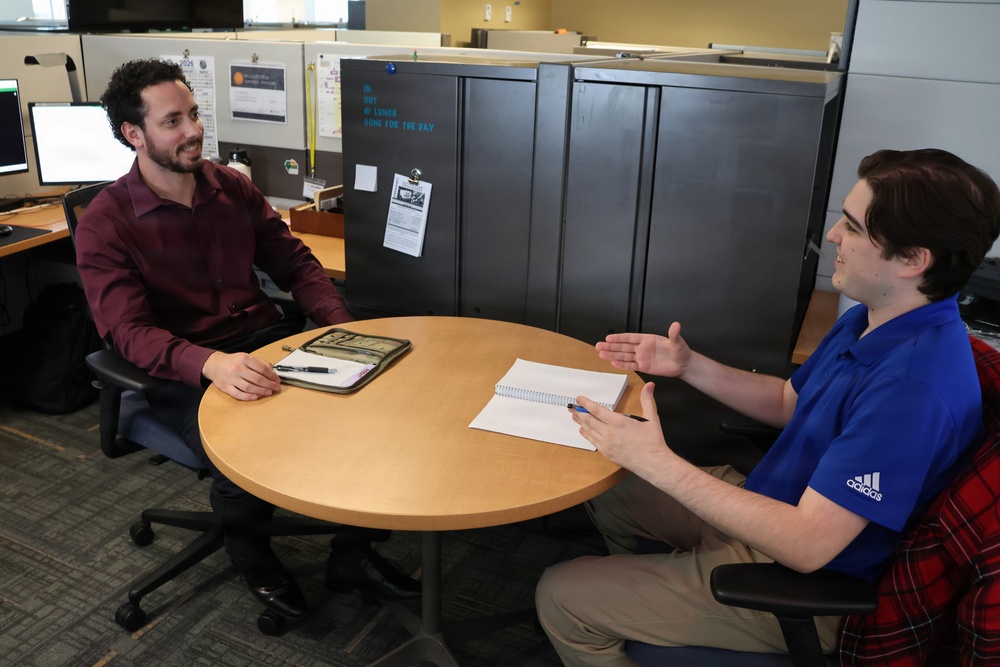 Two men are seated at a round wooden table in an open office environment, engaged in what appears to be a professional meeting or interview. The man on the right, wearing a blue Adidas polo shirt, is gesturing with his hand while holding a pen, suggesting he is actively speaking. The man on the left, in a dark red button-up shirt, is listening attentively with a slight smile.